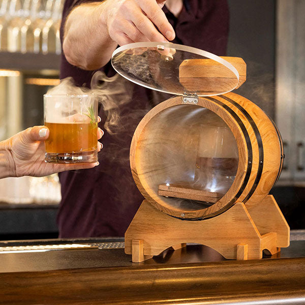 A person is using a cocktail smoker kit, which looks like a small oak barrel with a clear acrylic door, to smoke a cocktail. The smoker is placed on a bar counter.