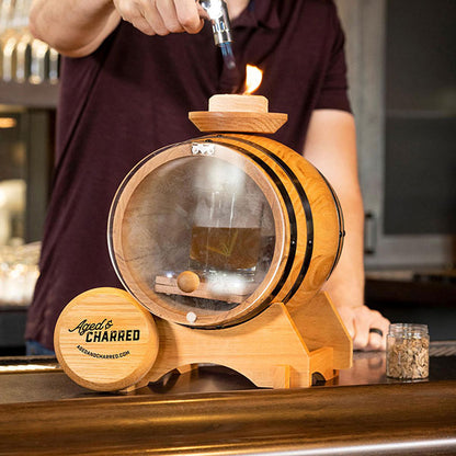 A person is smoking the glass of whiskey using the Aged & Charred smoke barrel kit that includes a 5-liter oak barrel, Smoke Lid, LED lights, wood chips, culinary torch, and butane.