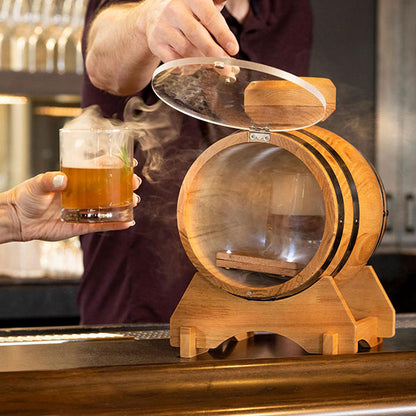 A person is using a cocktail smoker kit, which looks like a small oak barrel with a clear acrylic door, to smoke a cocktail. The smoker is placed on a bar counter.